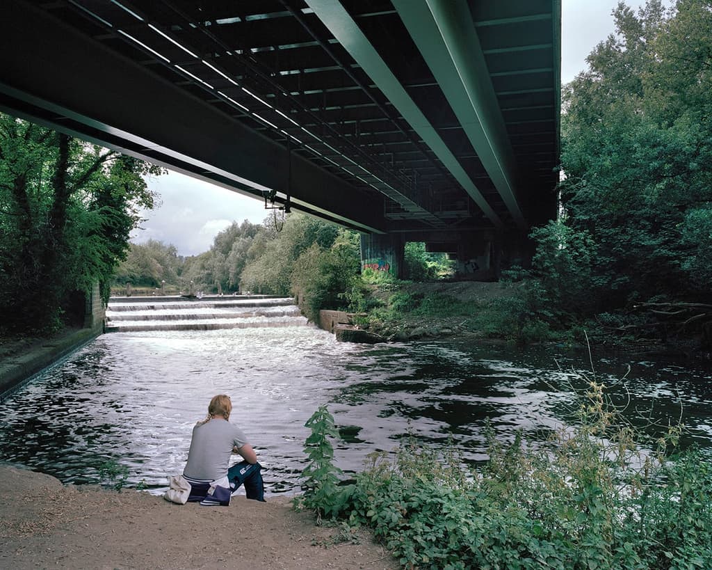 A lone man sits contemplating on the banks of the river, beneath the motorway pass