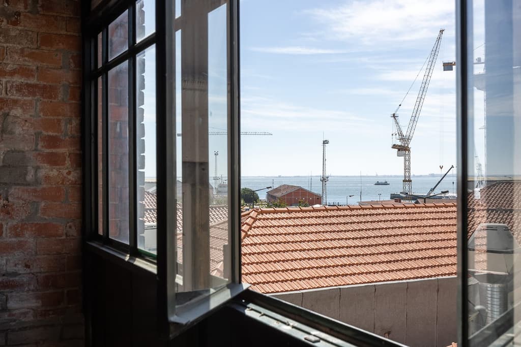 Patinated steel beams cross the space, supported by exposed brickwork while iron-framed windows overlook the Tejo estuary, shipyards beyond, and the city’s iconic Santuário de Cristo Rei in Almada.
