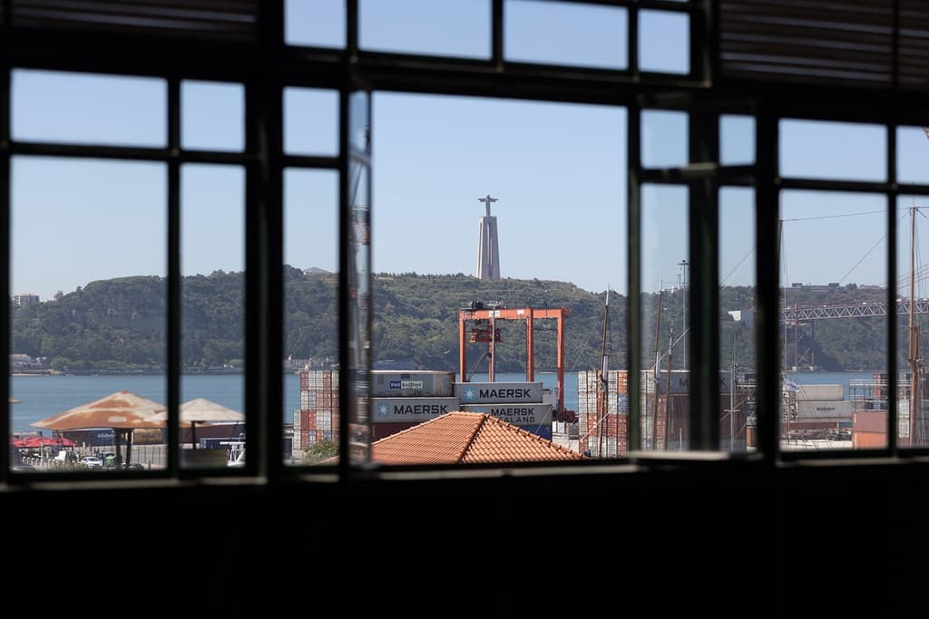 Patinated steel beams cross the space, supported by exposed brickwork while iron-framed windows overlook the Tejo estuary, shipyards beyond, and the city’s iconic Santuário de Cristo Rei in Almada.